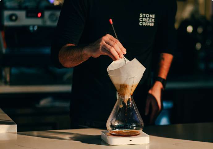 A person wearing a Stone Creek Coffee shirt stirs coffee grounds in a pour-over brewer on a scale.