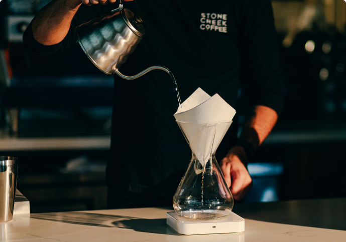 A barista pours water from a gooseneck kettle into a pour-over coffee maker on a scale.