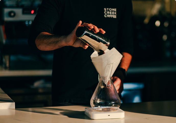 A Stone Creek Coffee barista pours coffee grounds into a paper filter in a glass pour-over brewer.