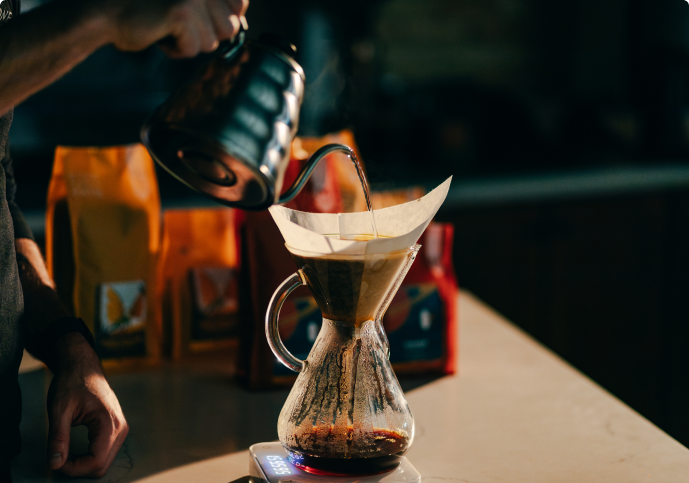 A person pours hot water from a gooseneck kettle into a glass pour-over coffee maker on a scale.