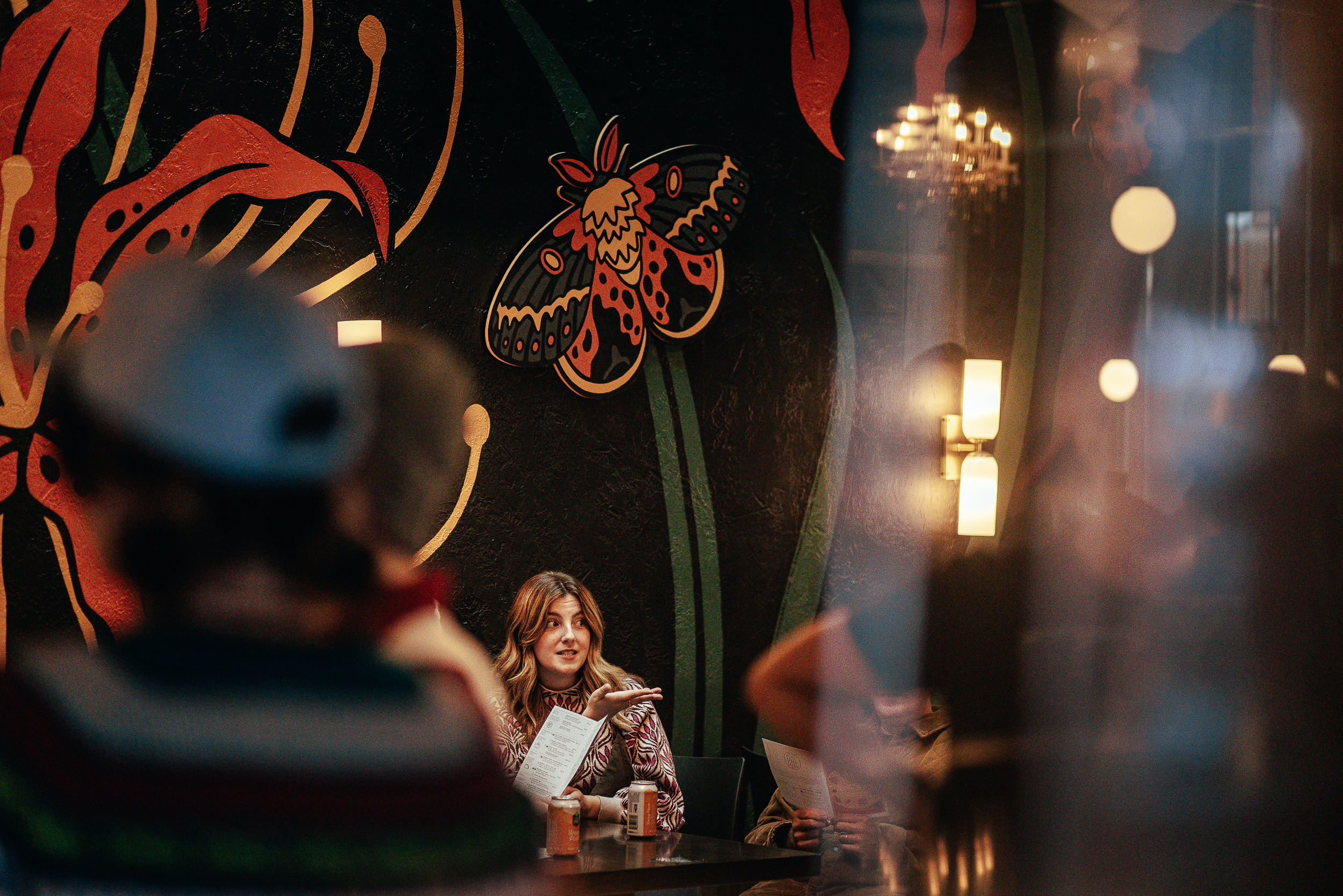 A woman gestures while holding a menu, seated at a table in front of a mural.