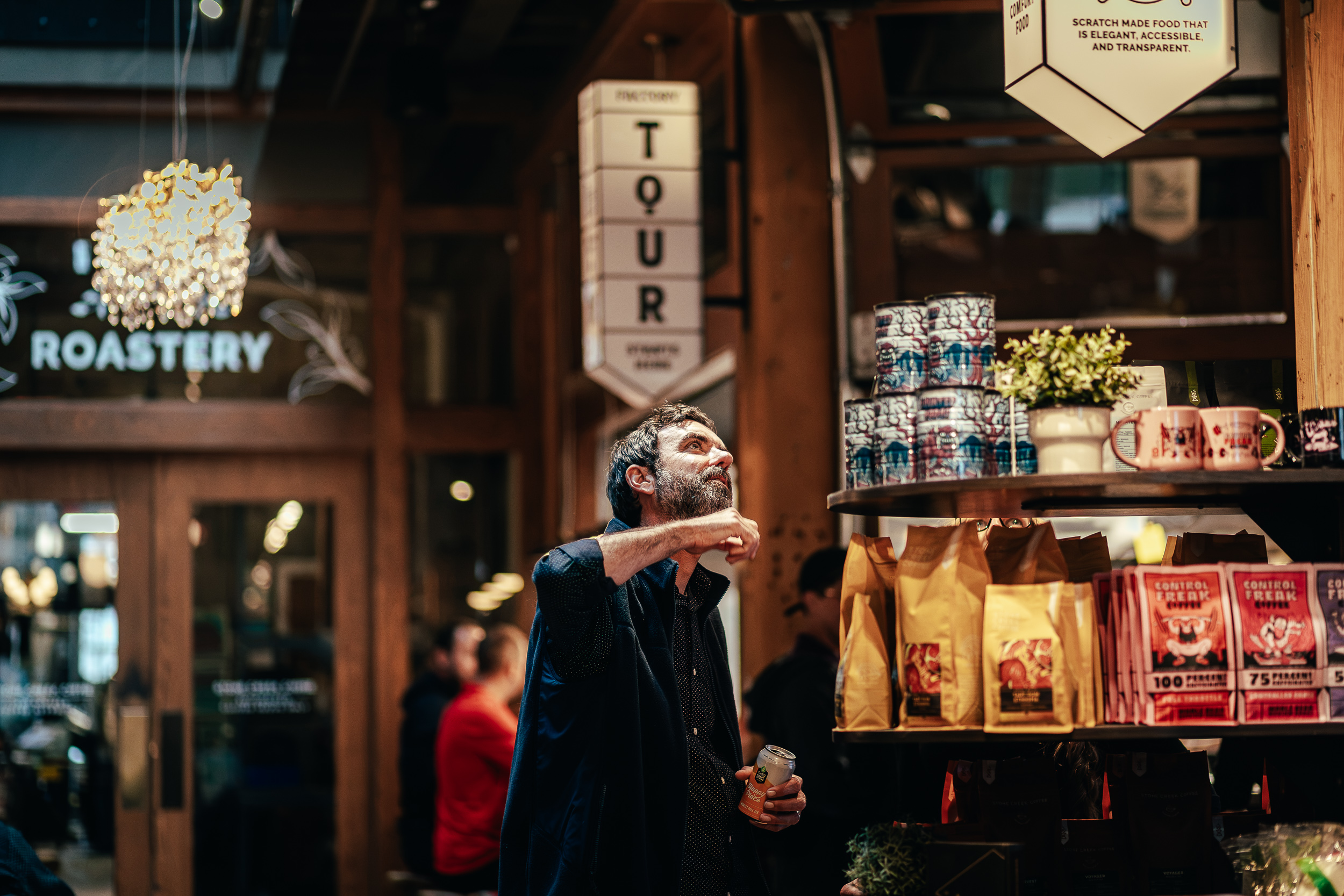 A bearded man looks up at products on shelves inside a busy market or roastery.