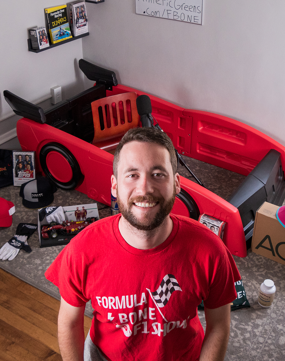 A smiling man in a red t-shirt sits in a room with a race car-shaped bed and memorabilia.