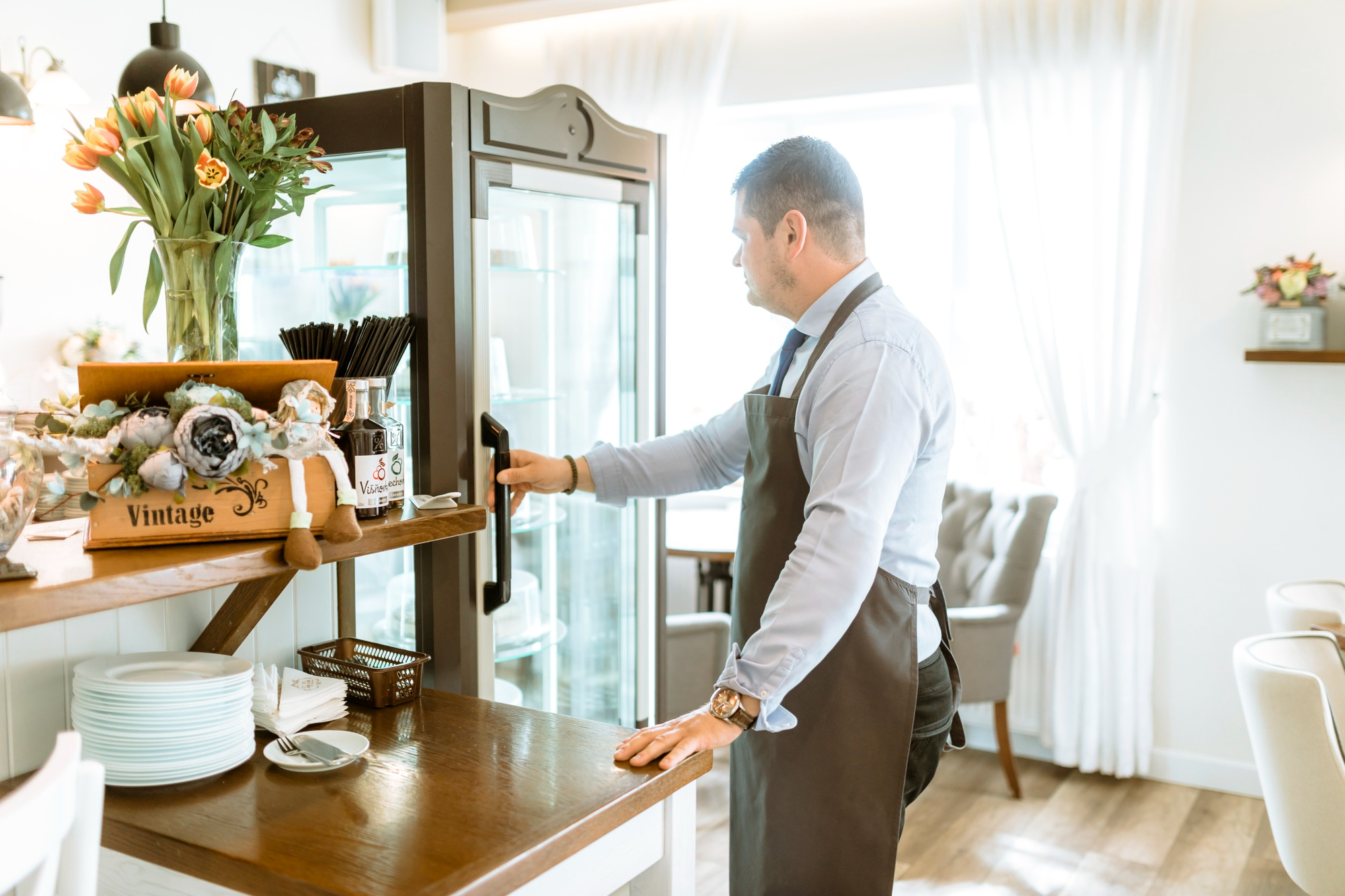 A man in an apron opens a glass display refrigerator in a brightly lit cafe or restaurant.