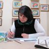 A woman with dark hair draws at a desk, with framed pictures on the wall behind her.