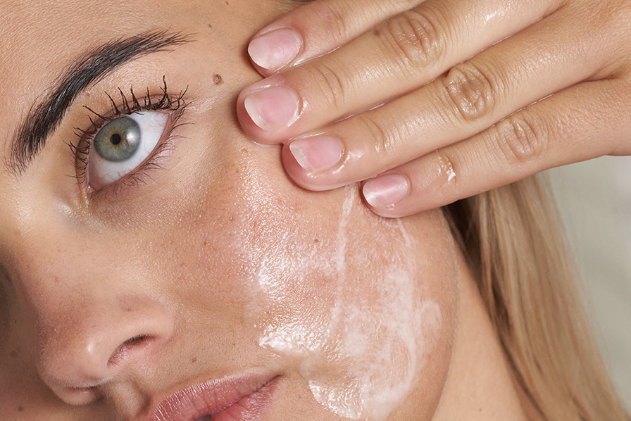 A close-up shot of a woman applying a white, foamy cleanser to her cheek with her hand.