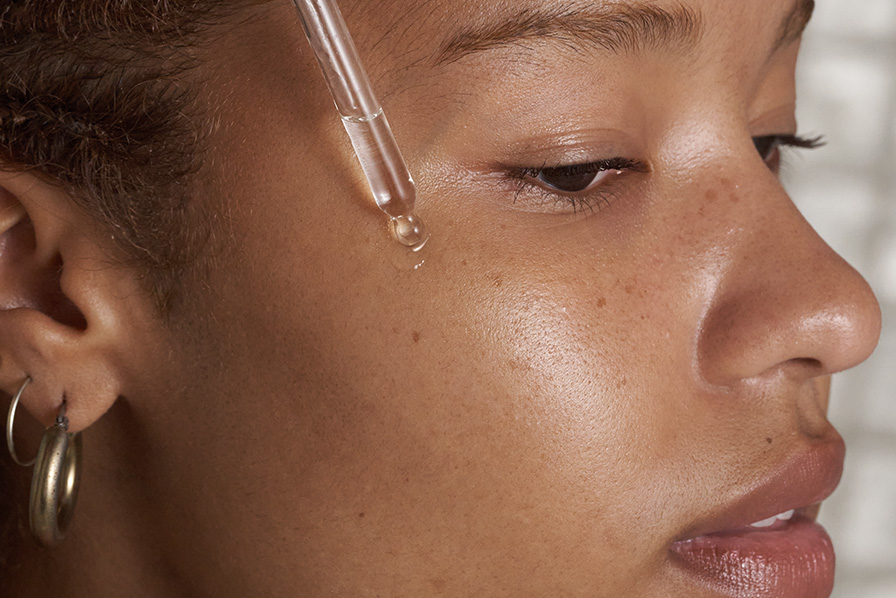 A close-up of a glass dropper applying a drop of clear liquid to a person's cheek.