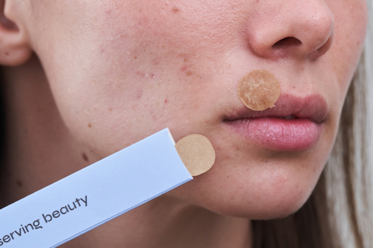 A close-up of a person's face with an acne patch, holding a packet of more patches.