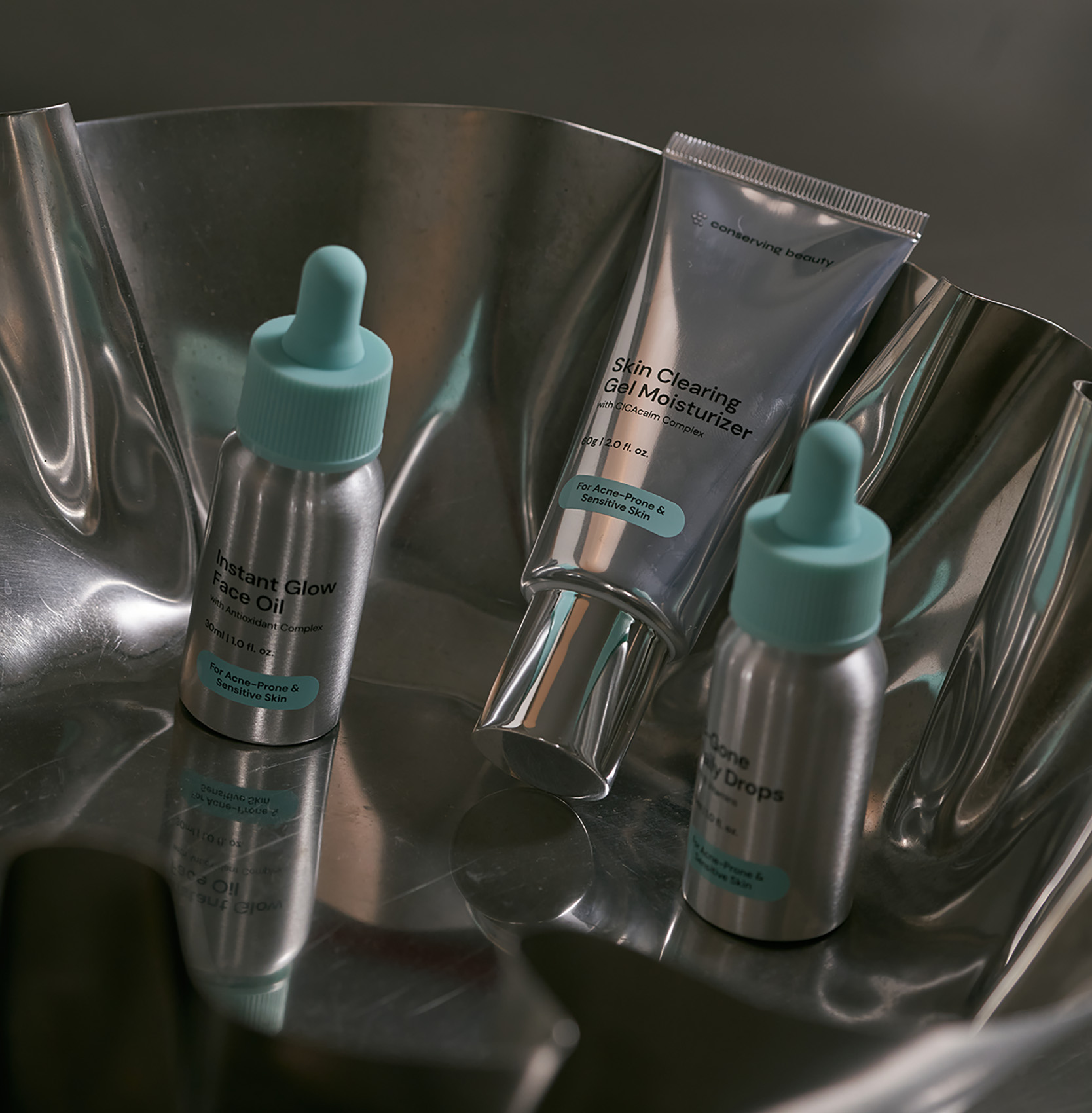 Three skincare products in silver packaging with mint green caps resting in a wavy, reflective metal bowl.