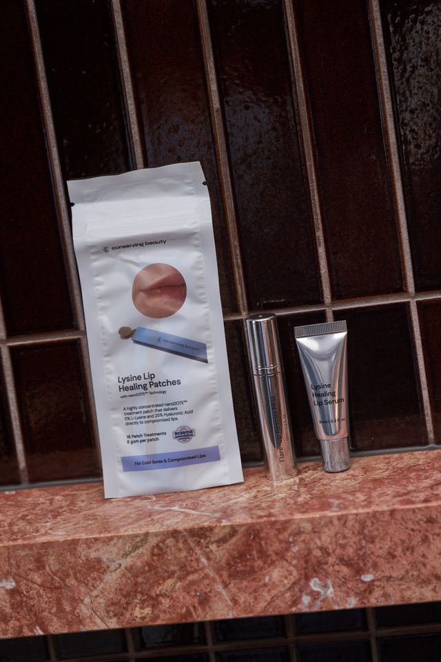 Conserving Beauty lip care products, including healing patches and serum, on a marble shelf against a tiled wall.