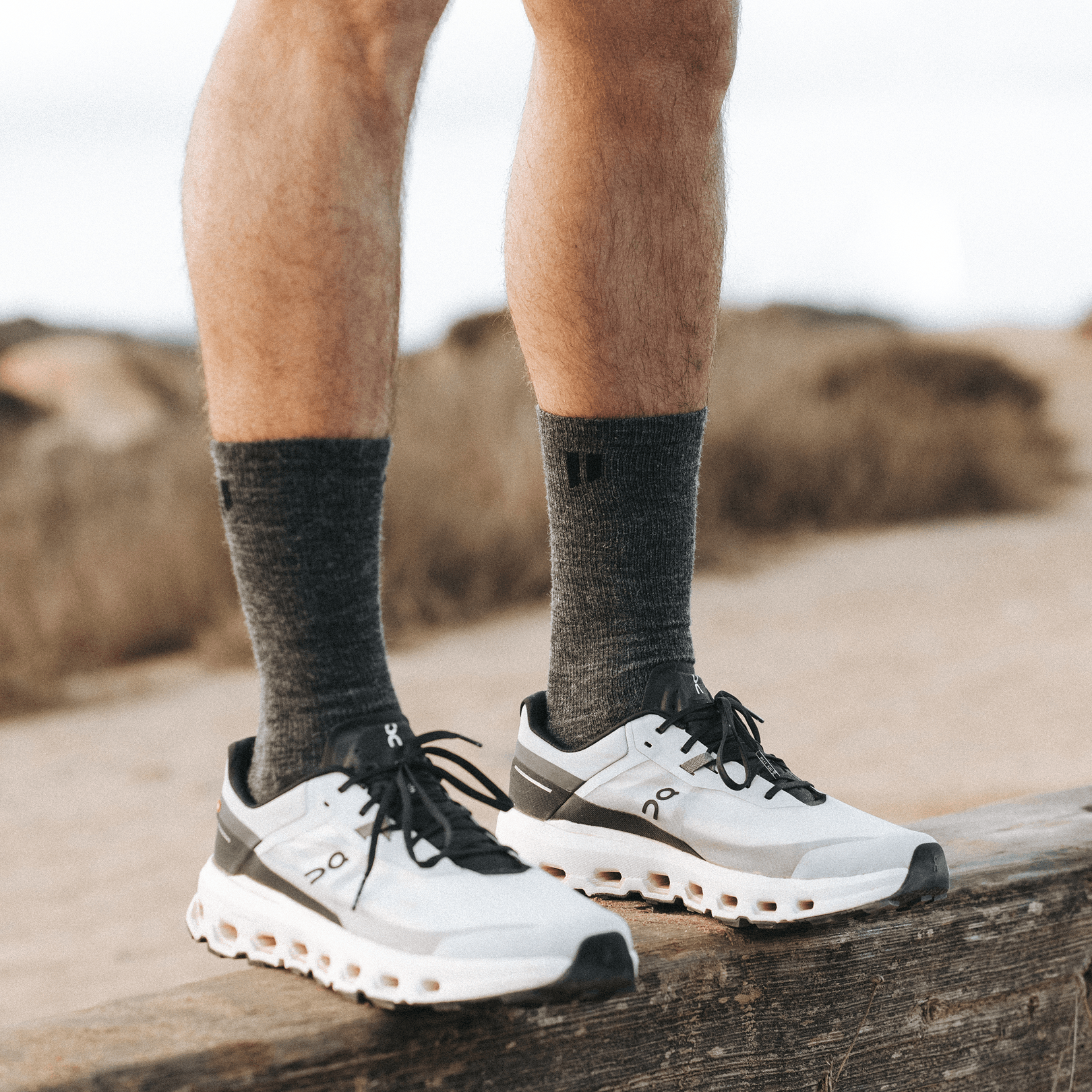 Close-up of a person's legs wearing grey socks and white running shoes, standing on a wooden beam.