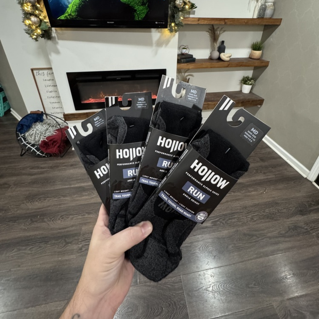A hand holding four new pairs of packaged black Hollow brand running socks indoors.