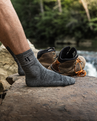 Person wearing gray sock next to hiking boots on a rock by a river.