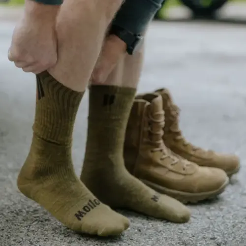 A person pulls on olive green socks with a pair of tan combat boots on the ground nearby.