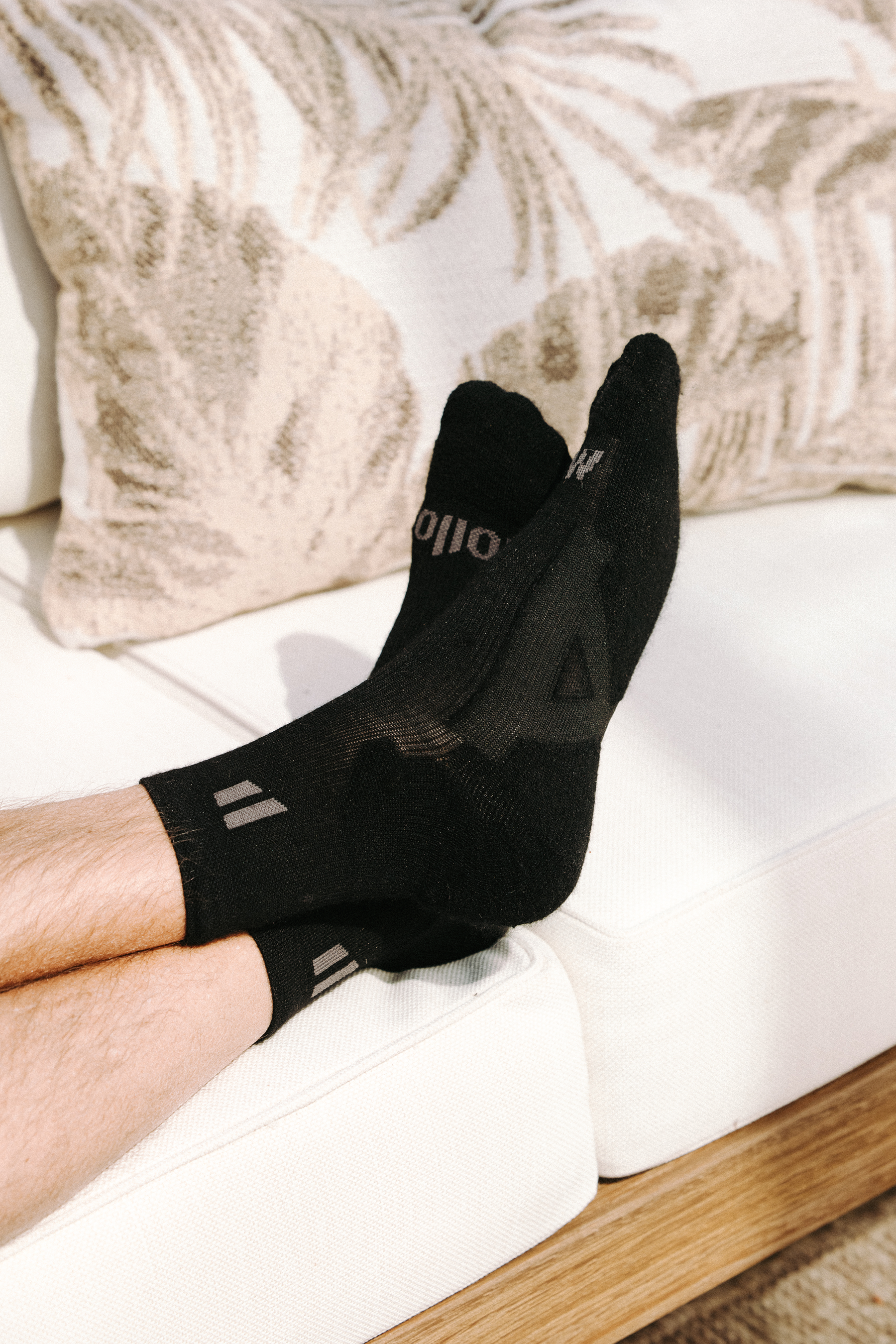A person's feet, wearing black socks, propped up on a white couch with a patterned pillow.
