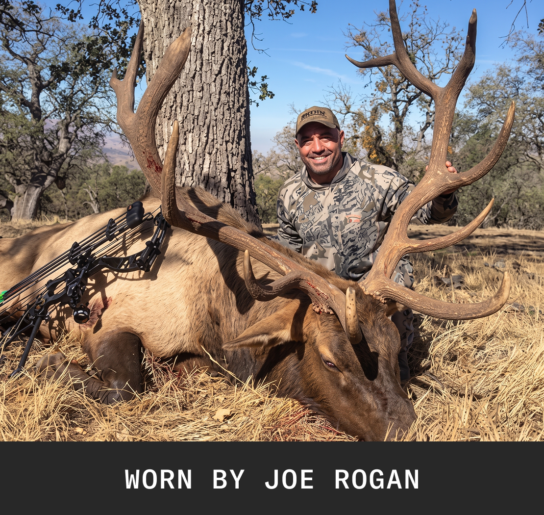 Joe Rogan in camouflage smiles while posing with a large elk he hunted with a bow.