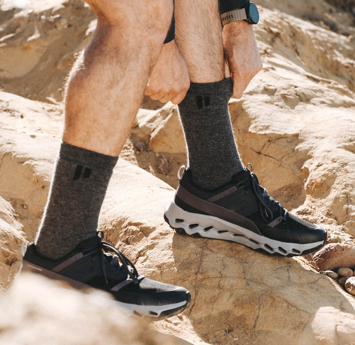 Man adjusting socks while hiking on rocky terrain wearing dark sneakers.