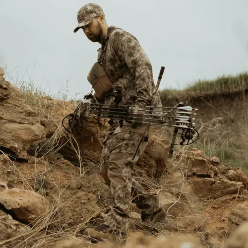 A man in full camouflage clothing carries a compound bow while walking up a rocky, grassy hillside.