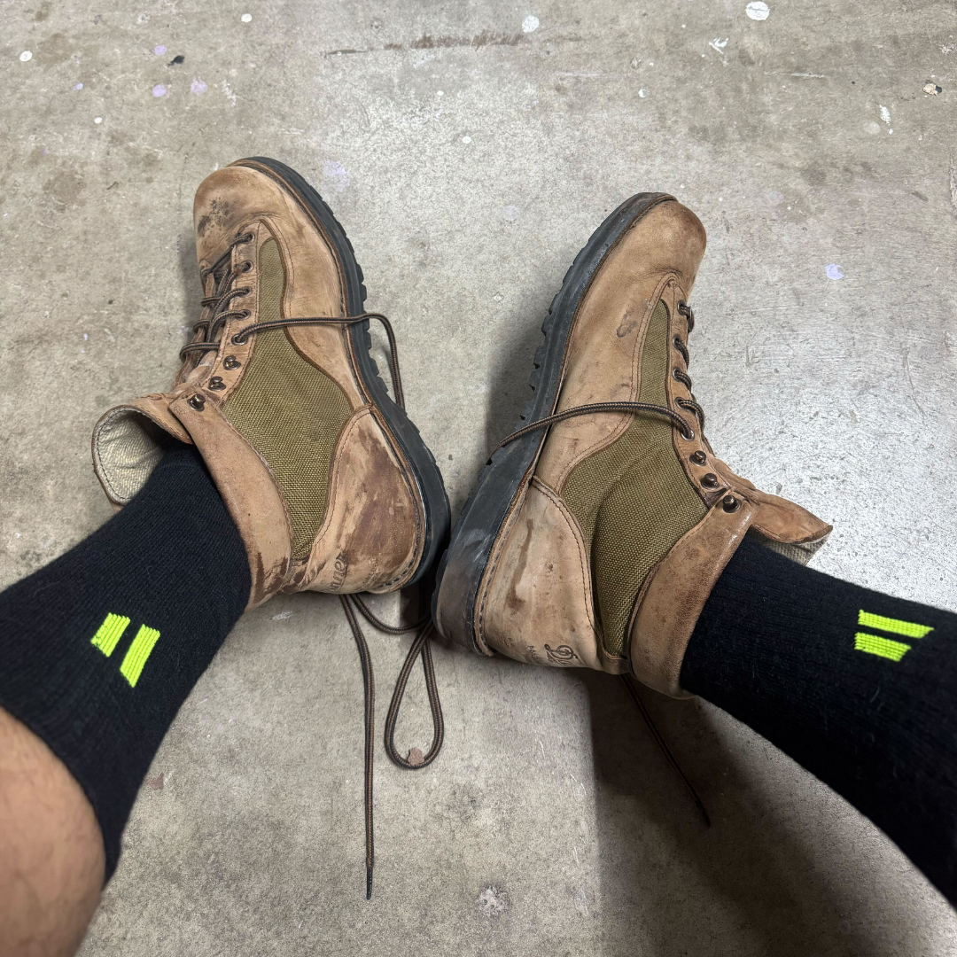 A first-person view of someone wearing muddy brown hiking boots and black socks on a concrete floor.