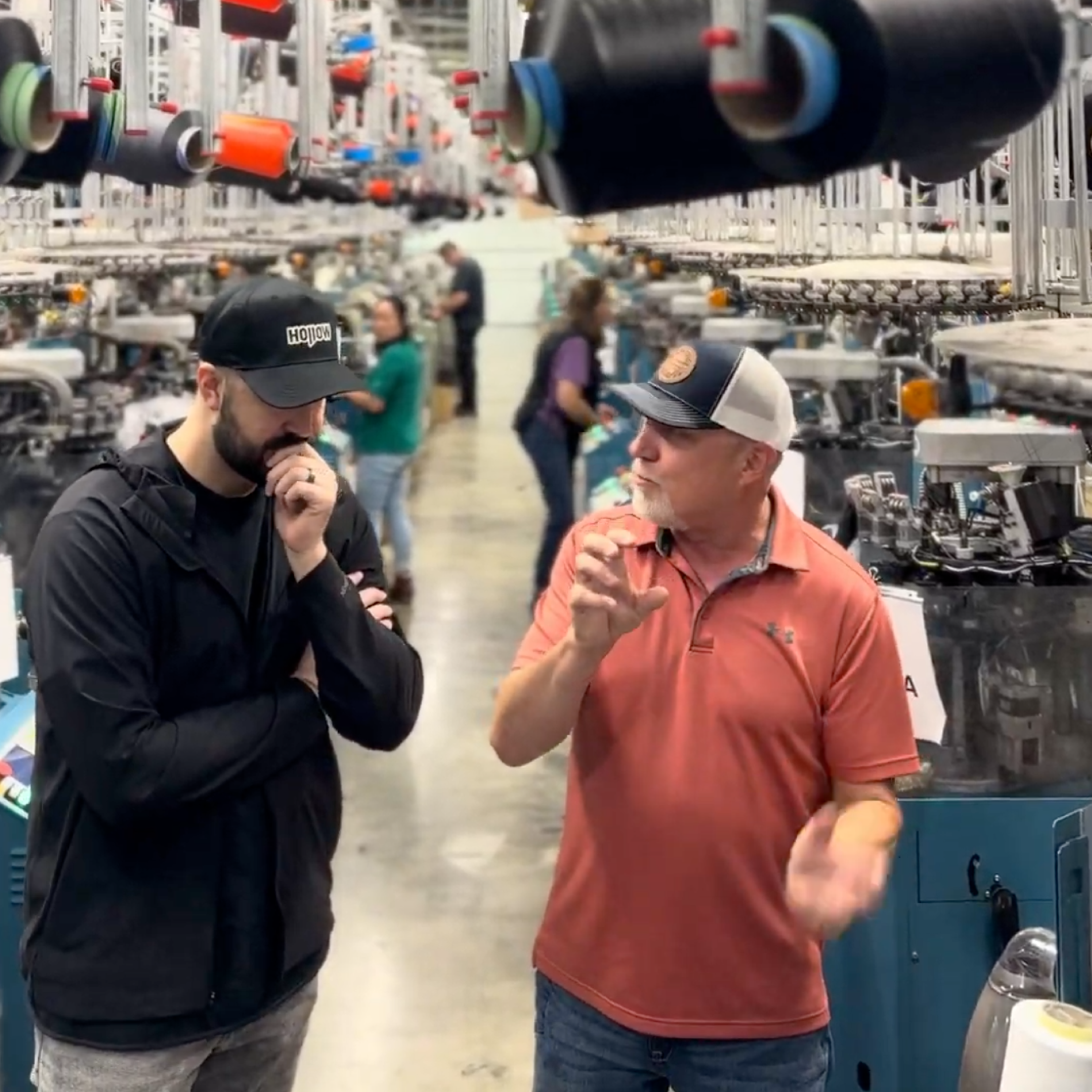 Two men talk in a textile factory, surrounded by machinery with large spools of black and red yarn.