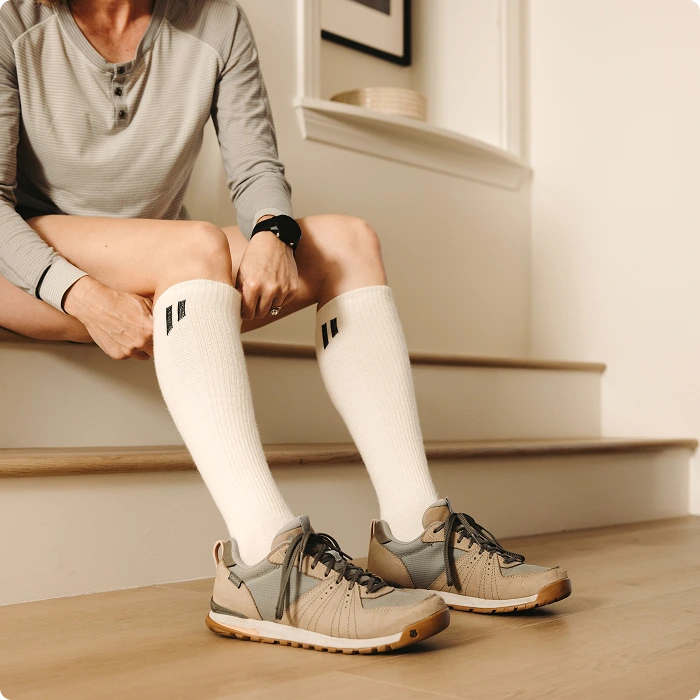 A person sits on wooden stairs wearing a grey shirt, white knee-high socks, and tan sneakers.