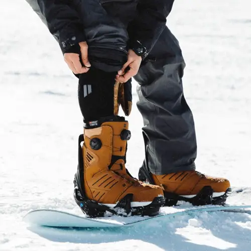 A person adjusts their snow pants over tan snowboard boots while standing on a snowboard in the snow.