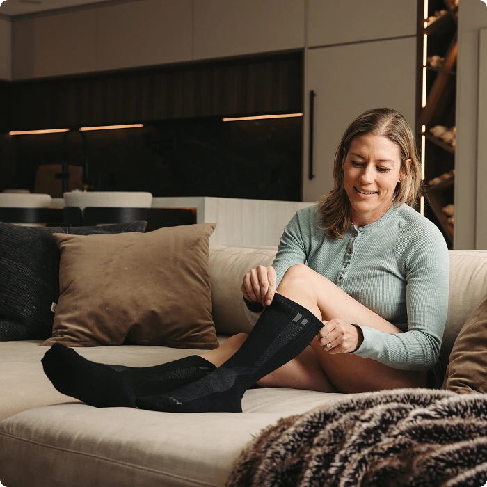 A woman sits on a couch putting on a pair of long black socks.