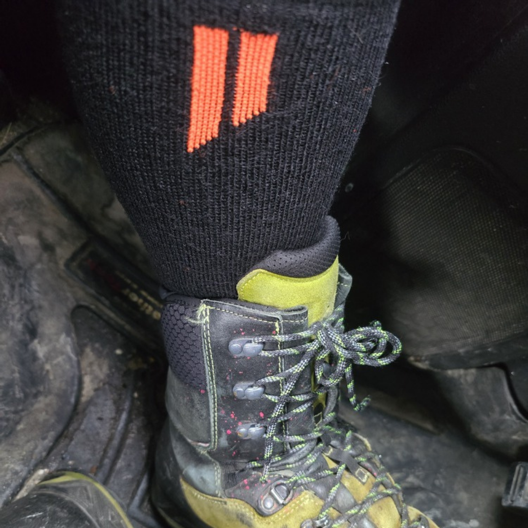 A close-up of a black sock with an orange logo worn with a sturdy, dirty green and black boot.