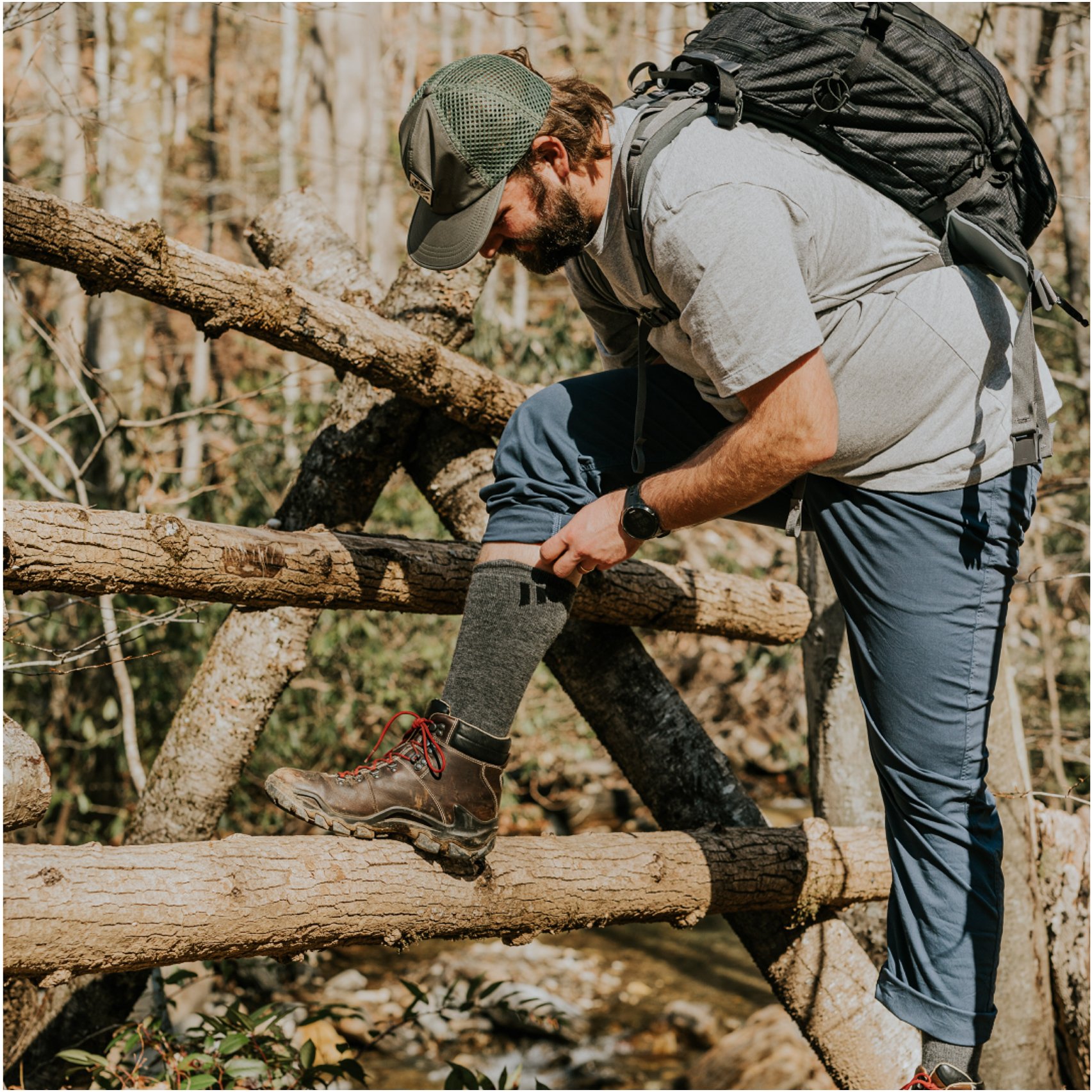Man adjusting sock while hiking on a wooden bridge.