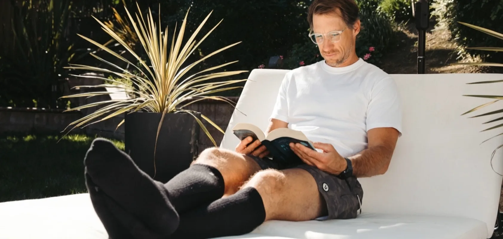 A man with glasses sits on a lounge chair in a garden, reading a book.