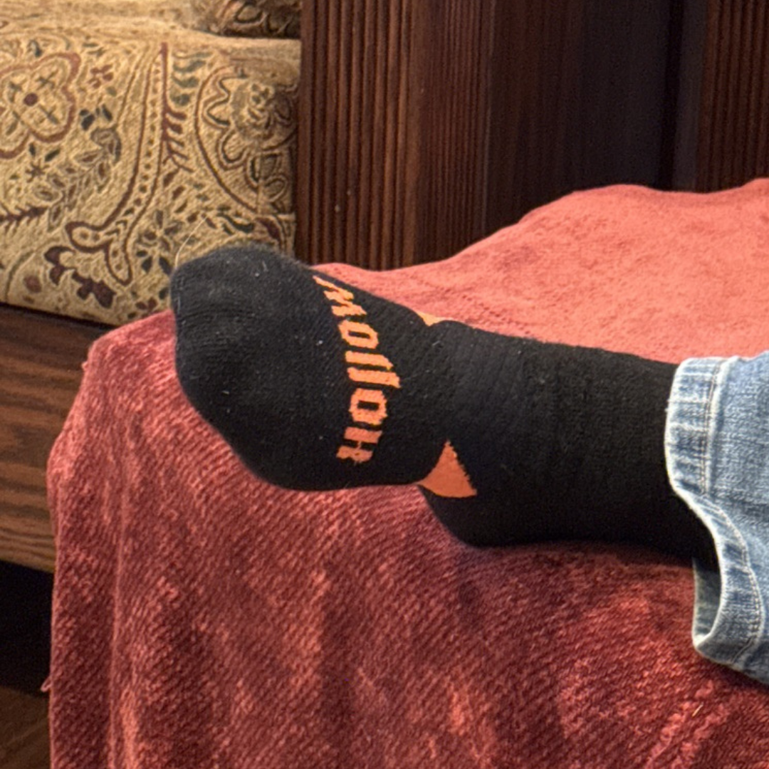 A close-up of a person's foot wearing a black sock with orange lettering, resting on a red blanket.