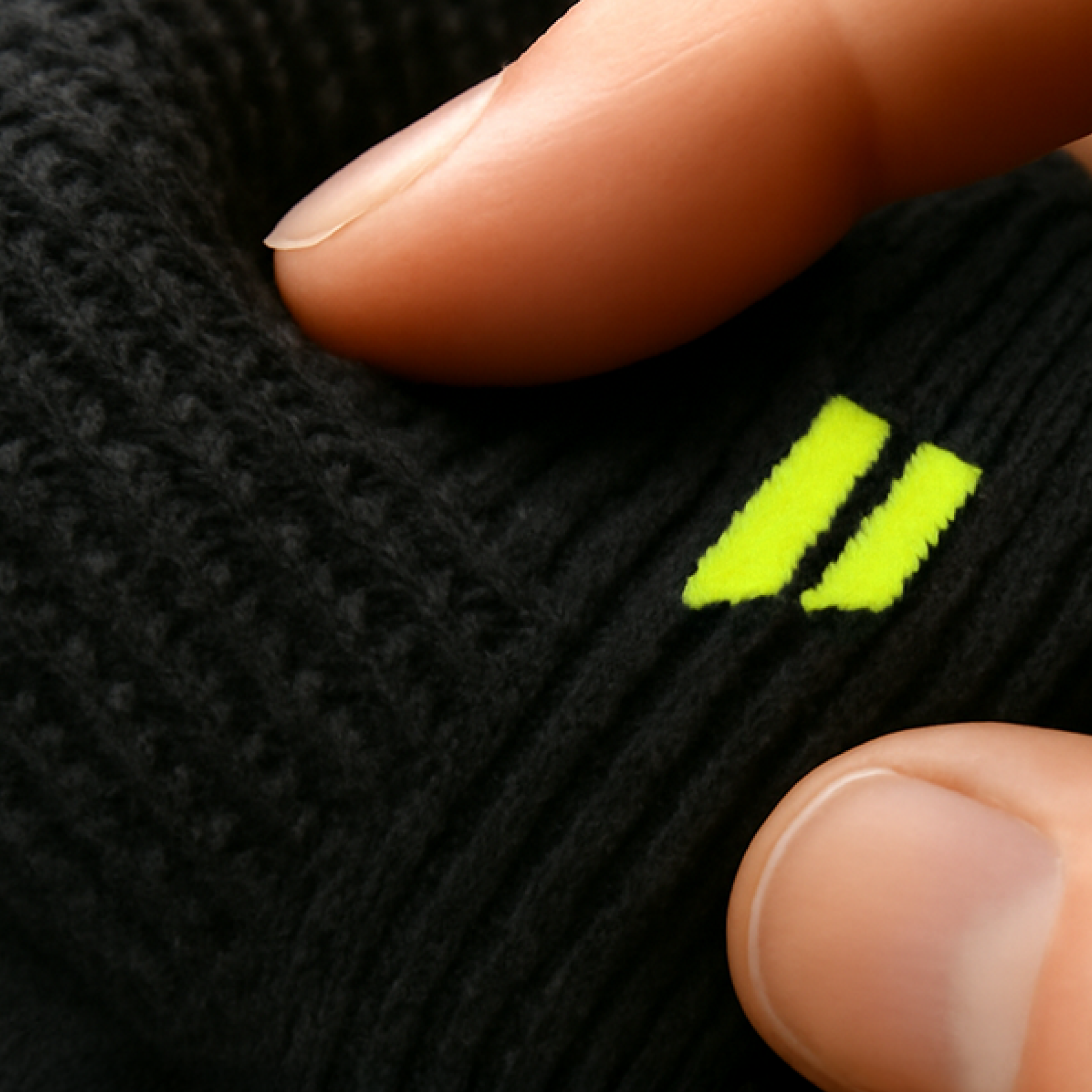 A close-up of fingers pinching a black, ribbed fabric with a small, neon yellow two-stripe logo.
