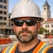 A bearded man wearing a white hard hat, sunglasses, and a high-visibility vest outdoors.