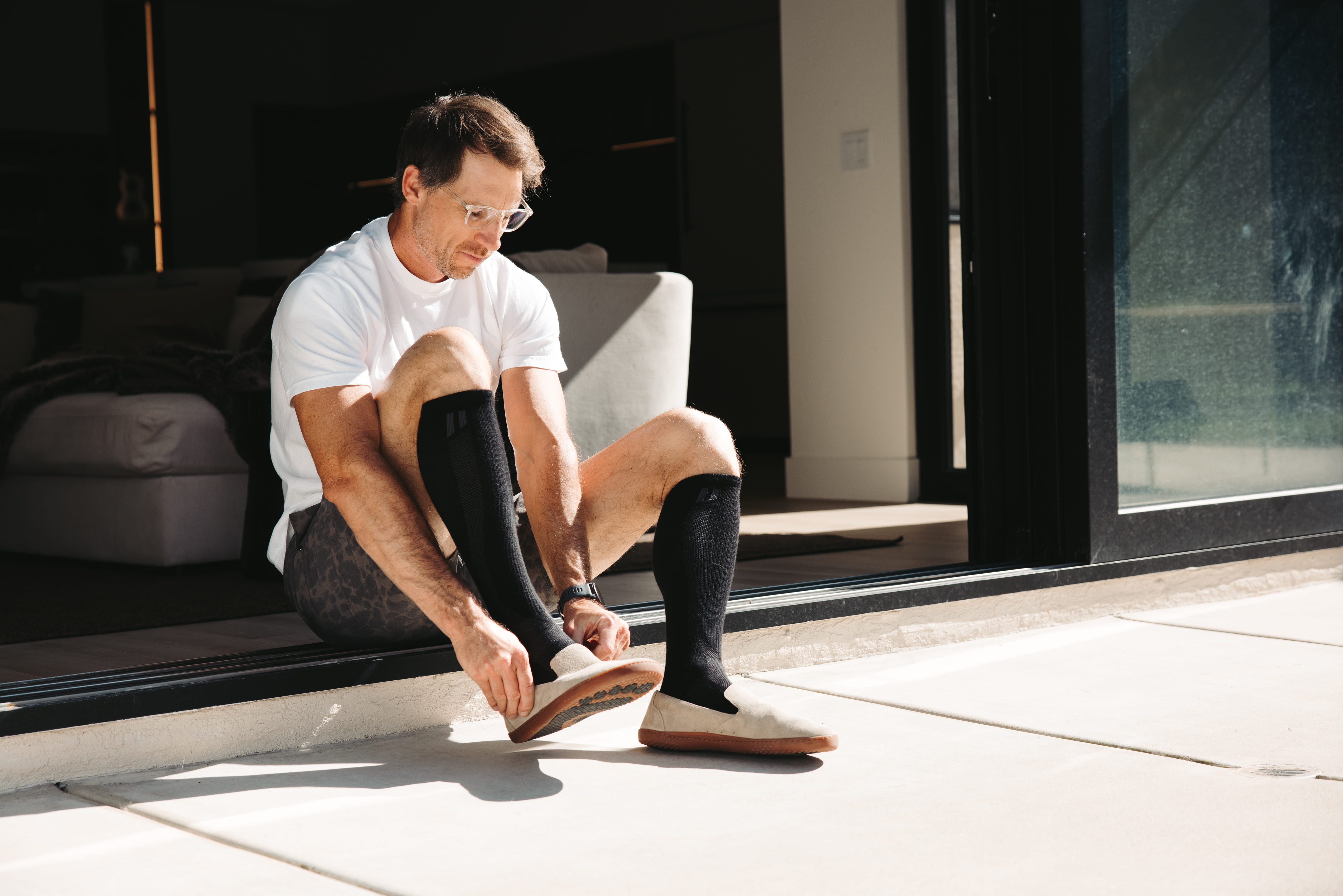 A man wearing glasses and black socks sits in a sunny doorway putting on a slip-on shoe.
