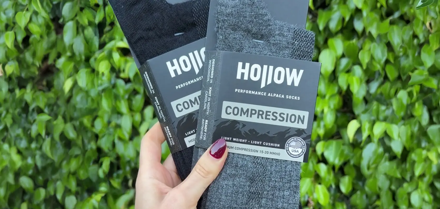 A hand holds two pairs of black and grey Hollow brand compression socks against a leafy green background.