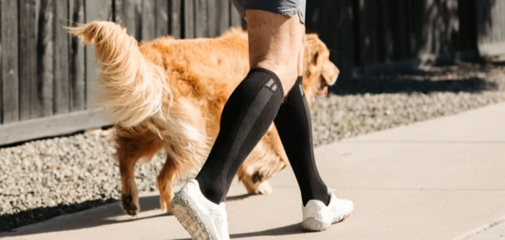 A person wearing black compression socks and white sneakers walks a golden retriever on a sidewalk.