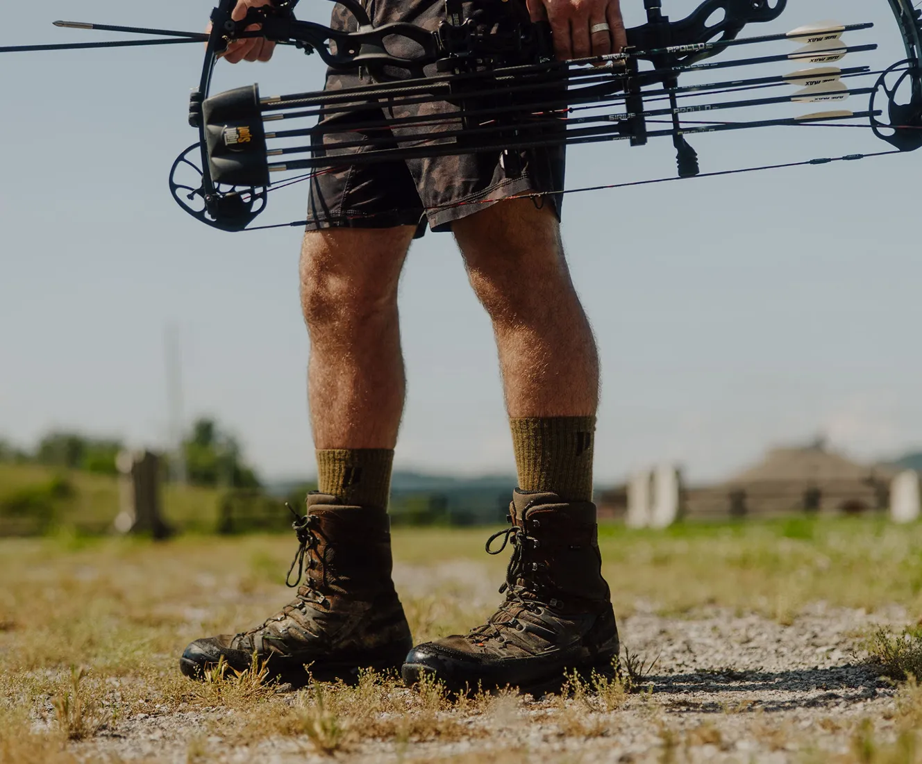 A person wearing camouflage shorts and hunting boots holds a compound bow in a grassy field.