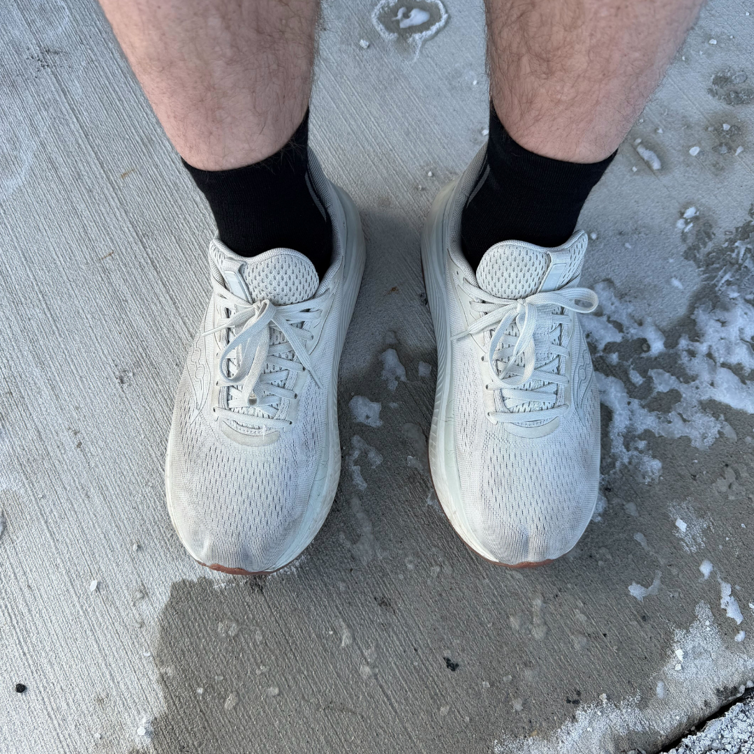 A person's feet in white running shoes and black socks standing on a snowy concrete sidewalk.