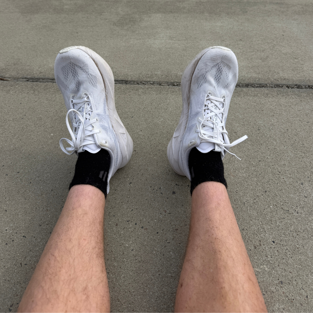 A first-person view of a person's legs wearing white running shoes and black socks on pavement.