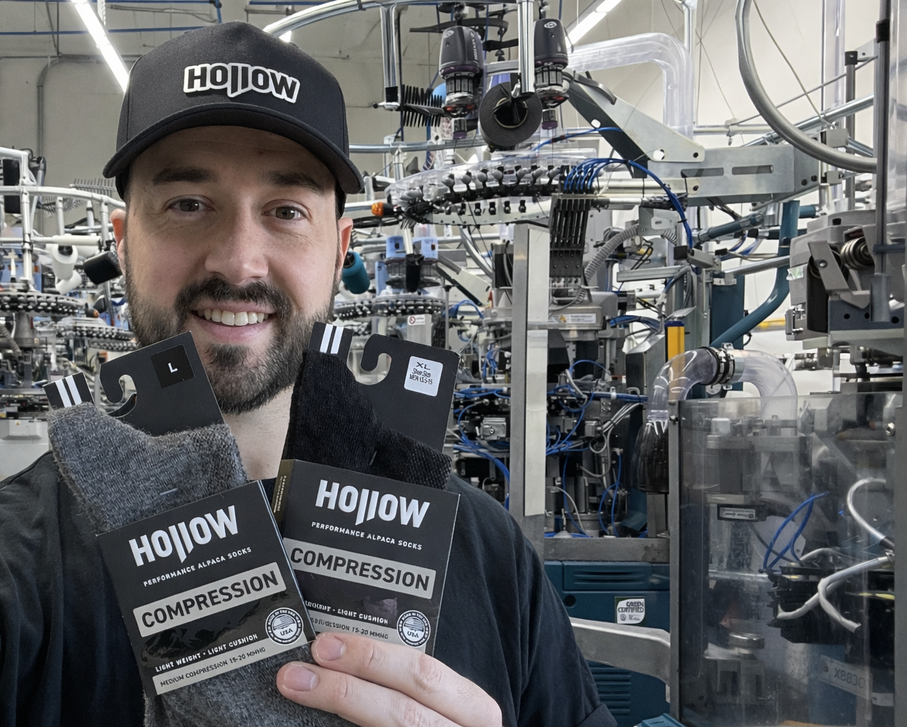 A man in a hat holds two pairs of Hollow brand compression socks in a factory.