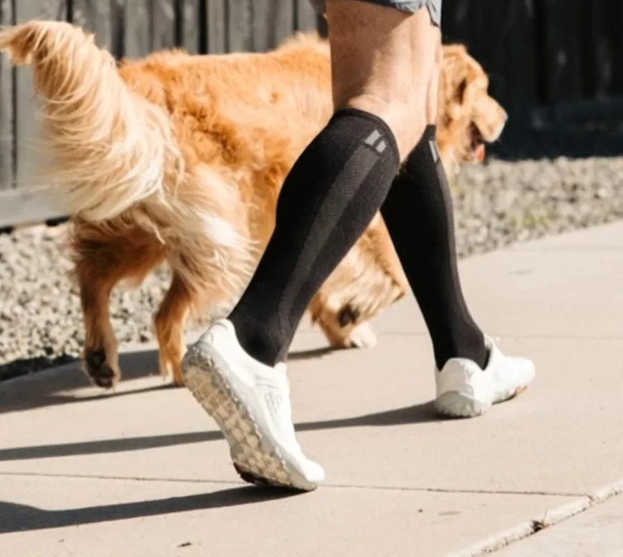A person wearing black compression socks and white sneakers walks a golden retriever on a sidewalk.
