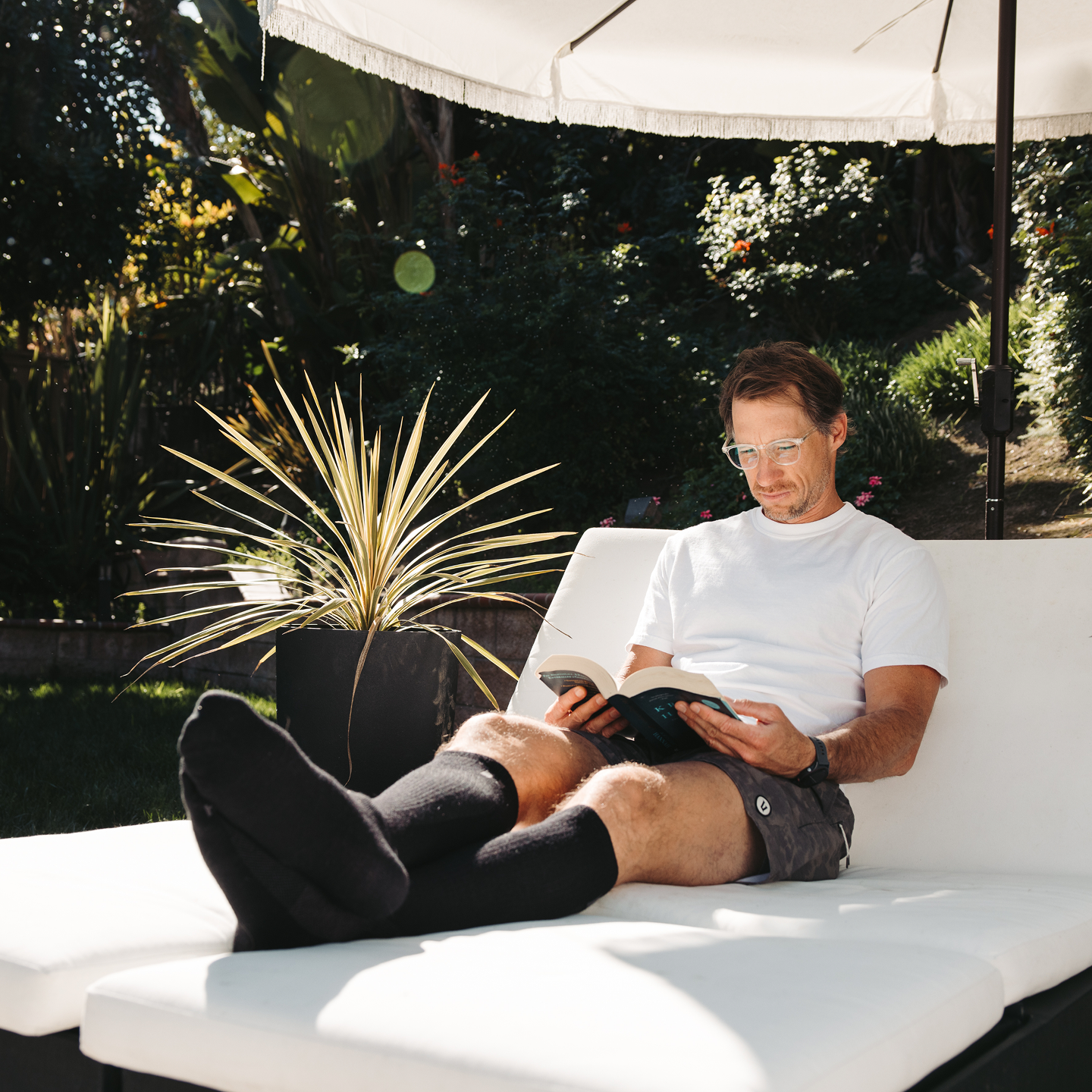 A man wearing glasses reads a book while relaxing on a white lounge chair outdoors.