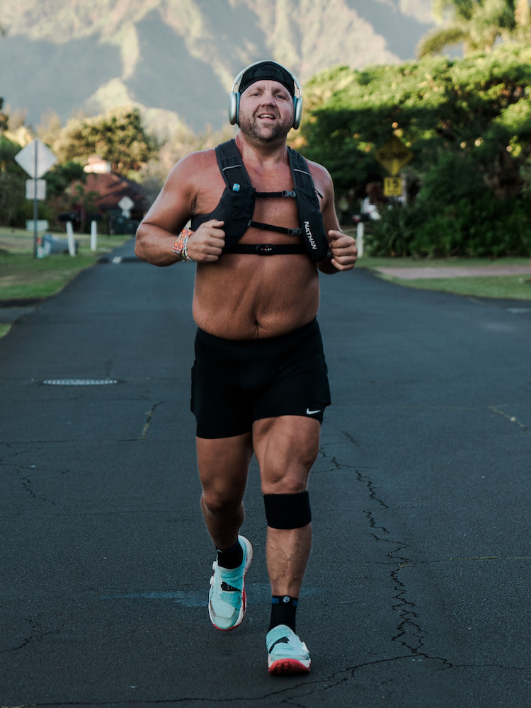 Man jogging on a road with headphones and knee brace, mountains in background.