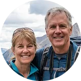A smiling man and woman wearing backpacks, with mountains in the background.