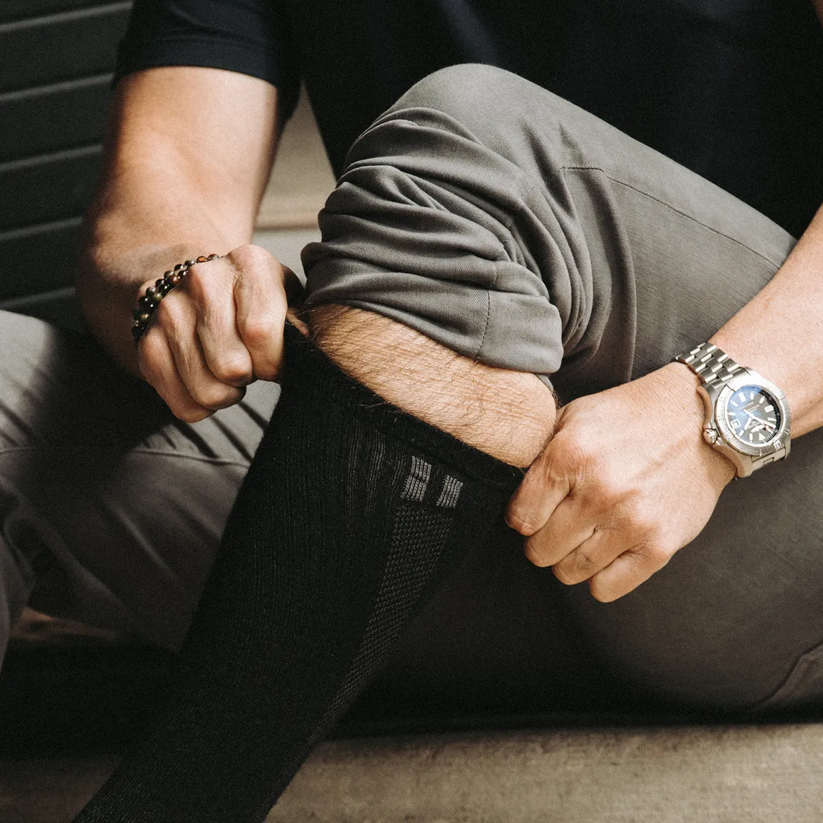 A man in grey trousers sits down while pulling on a long black sock.