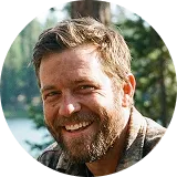 A circular headshot of a smiling man with a beard, outdoors.