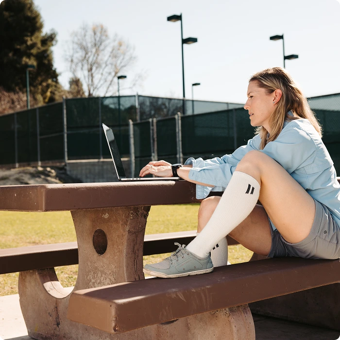 A woman sits at an outdoor picnic table, wearing tall socks and working on a laptop.