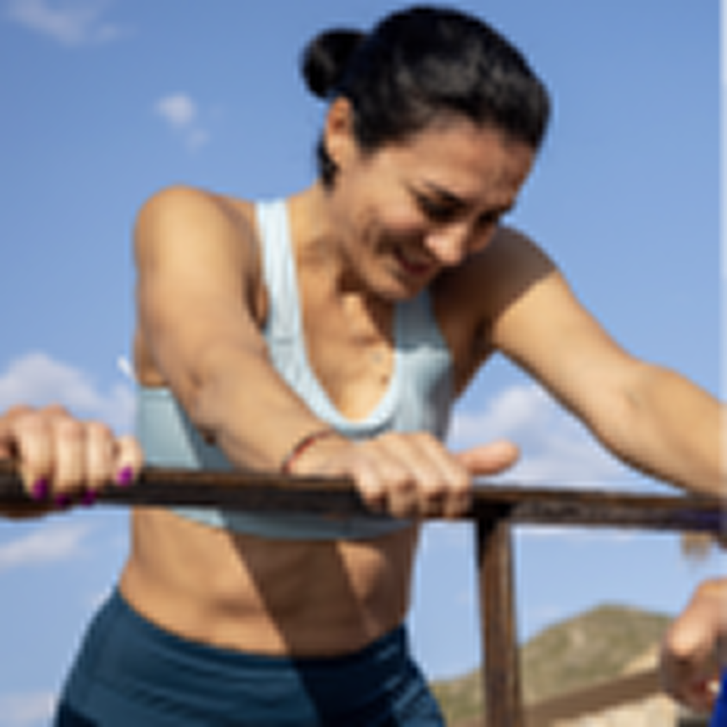 A woman in athletic wear grimaces as she leans over a bar during an outdoor workout.