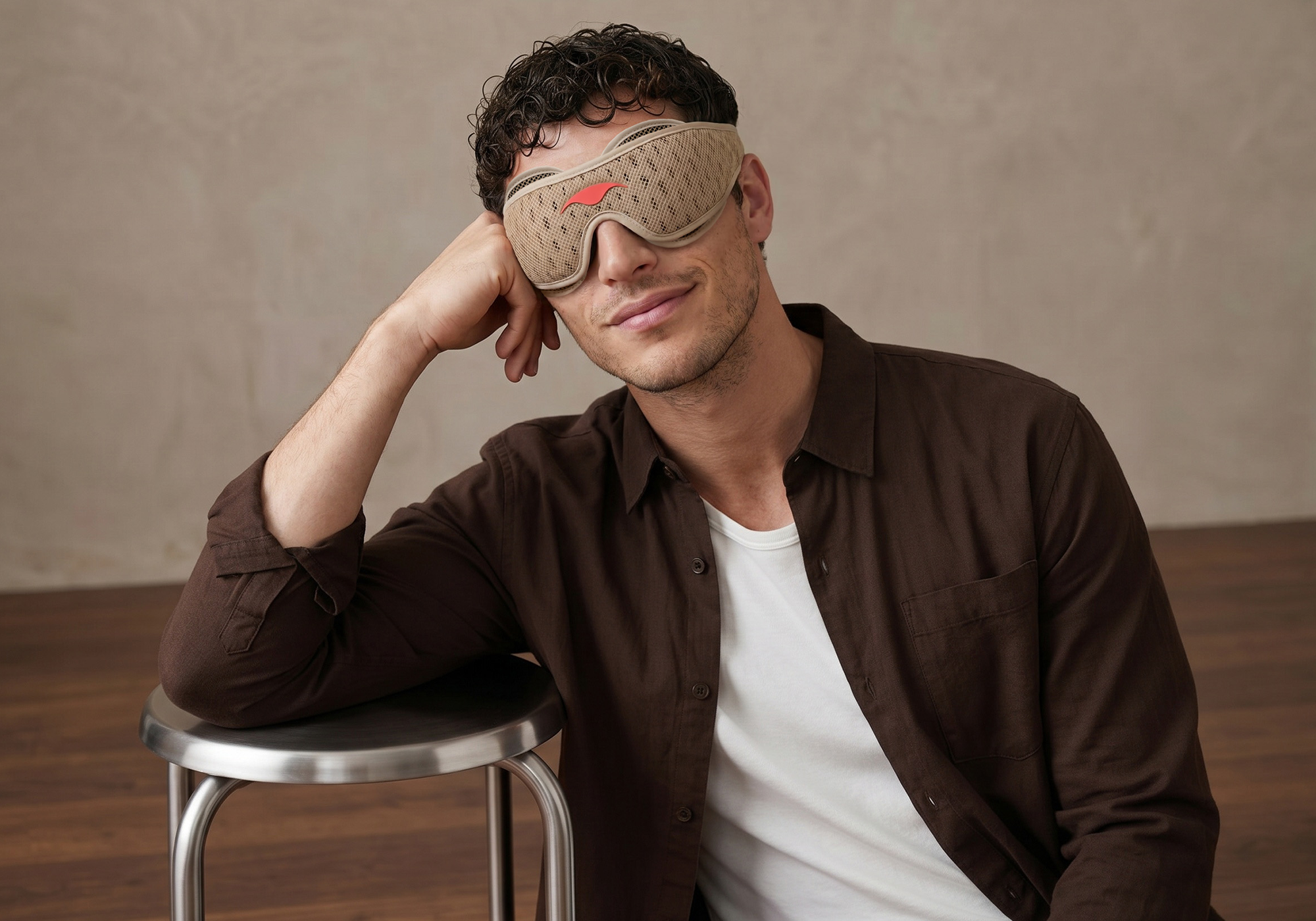 A man wearing a tan sleep mask sits on a stool and smiles at the camera.
