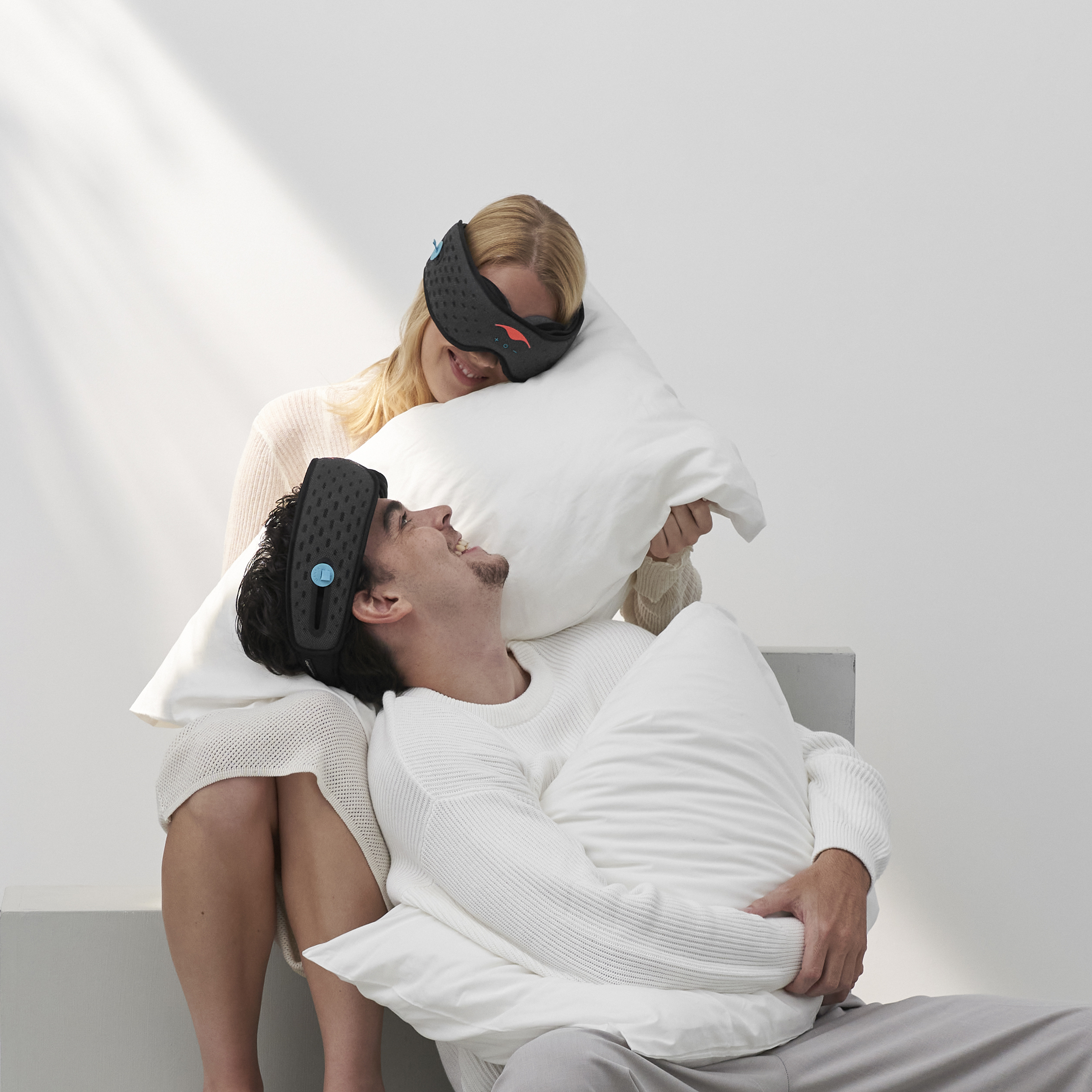 Two people wearing eye masks, holding pillows, and smiling at each other.
